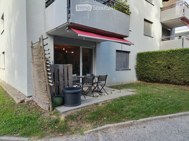 Outdoor patio area with a small table, chairs, and potted plants, adjacent to a modern building with balconies and a red awning.