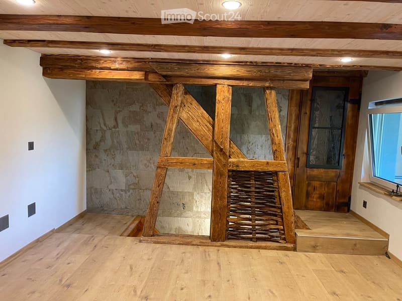 Empty room with wooden floor, exposed wooden beams, and a stone wall. Features a wooden door with decorative glass panel and a window with a shelf.