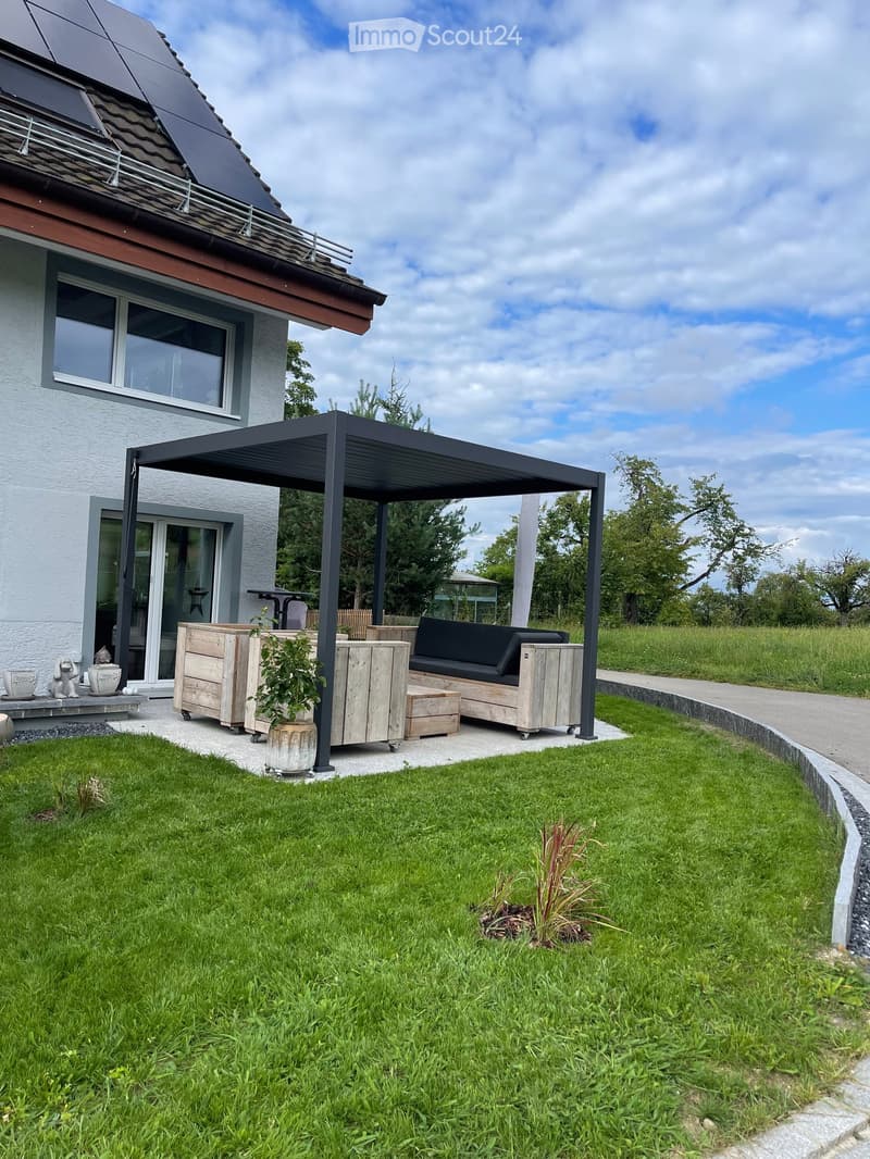 Outdoor patio area with a black metal pergola, wooden furniture including a sofa, table, and chairs, and potted plants. The area is surrounded by a well-maintained lawn and a modern house with large windows and a solar panel on the roof.