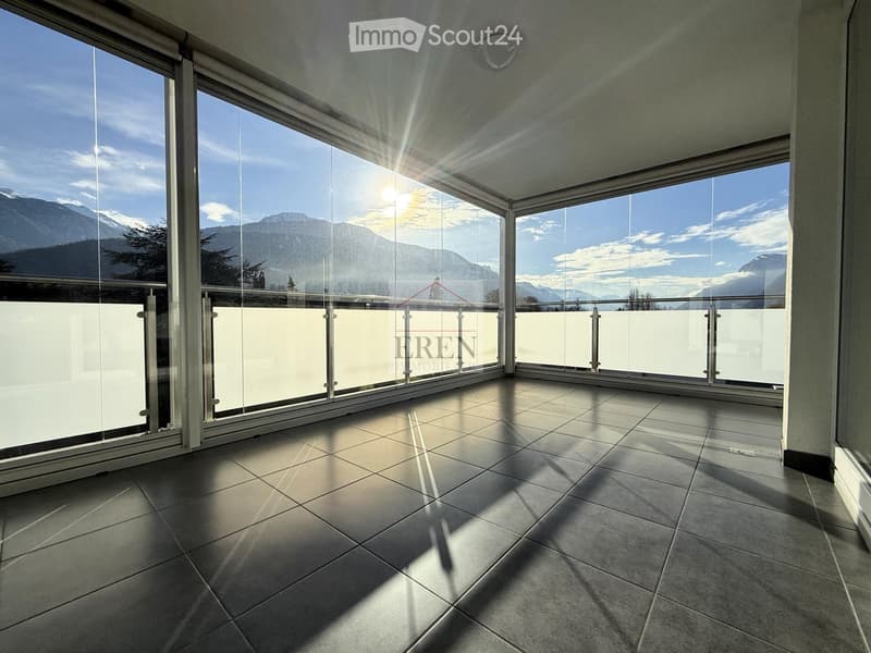 Spacious balcony with large glass windows and a modern glass railing, offering a scenic mountain view.
