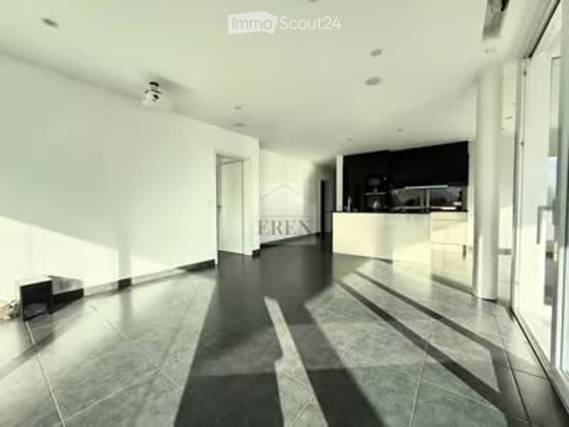 Bright, empty room with white walls, dark tiled floor, and a small kitchen area visible in the background. No furniture or personal items present.