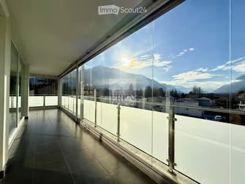 Spacious balcony with large glass windows and a modern glass railing, offering a scenic mountain view.
