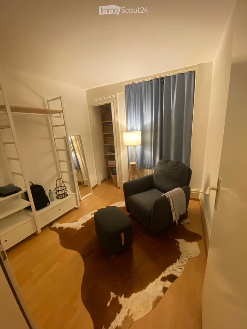 A cozy living room with a dark grey sofa, a black ottoman, a floor lamp, a white shelf, and a large mirror. The room features wooden flooring and a cowhide rug.