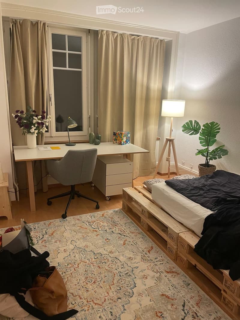 A cozy, well-lit room with a bed, desk, and various decorative items. The space features a large window with curtains, a patterned rug, and a mix of modern and rustic furniture.