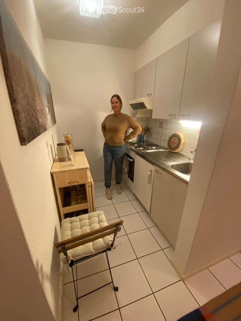 A small, well-lit kitchen with white tiled flooring, white cabinets, a sink, and a small wooden table with a chair. A person stands in the center, and a painting is visible on the wall.