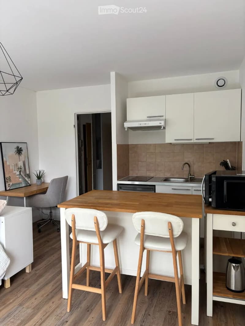 Modern kitchen with white cabinets, wooden island, bar stools, microwave, sink, and a small workspace area with a chair and desk.