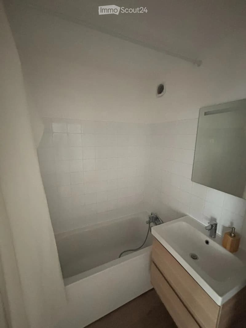 Bathroom with a bathtub, sink, mirror, and wooden cabinet. The walls are tiled, and there is a small window providing natural light.