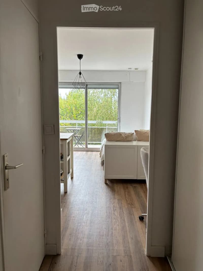 Modern, minimalist room with light wooden flooring, white walls, and a large window leading to a balcony. Visible furniture includes a sofa, a small table, and a pendant light.