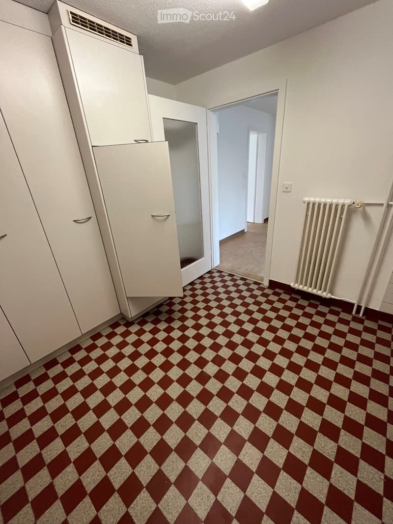 Empty room with white walls, checkered red and beige floor, white radiator, and white cabinet with mirrored door.