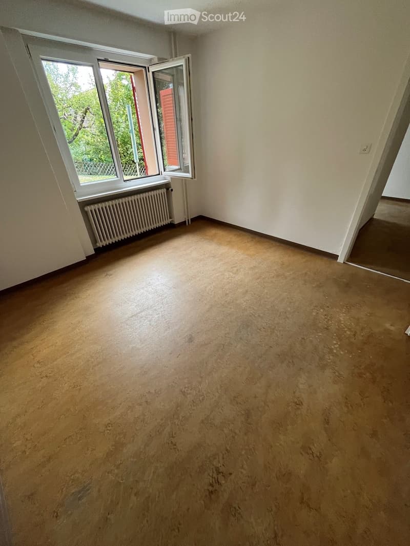 Empty room with light-colored wooden flooring, white walls, and a radiator beneath a large window with open shutters.