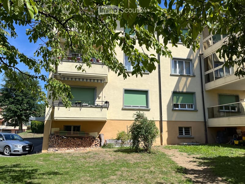 Four-story residential building with balconies, green shutters, and a small garden area with a stack of firewood and a parked car.