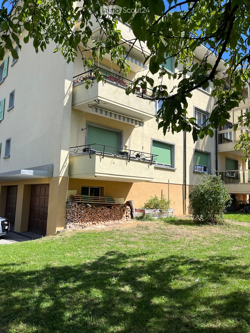 A multi-story residential building with balconies, green shutters, and a neatly maintained lawn in front. A stack of firewood is visible near the entrance, and a car is parked nearby.