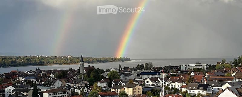 A panoramic view of a town with houses, a church tower, and a rainbow over a body of water.
