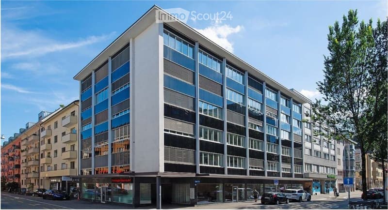 Modern multi-story building with large glass windows and balconies, located in an urban setting with surrounding structures and parked cars.