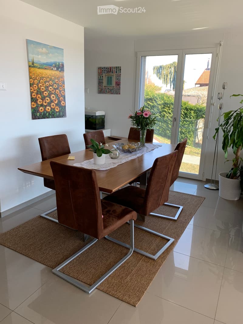 Dining room with a wooden table, six brown chairs, a vase with flowers, a small plant, and artwork on the walls. The room has large windows and a view of greenery outside.