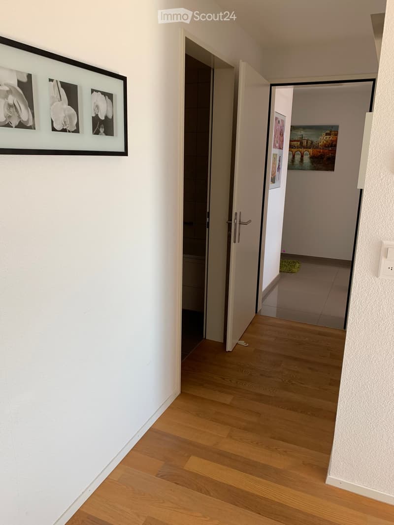 A clean, well-lit hallway with wooden flooring, white walls, and several open doors leading to other rooms. Artwork is displayed on the walls, and a small green rug is visible in one of the adjacent rooms.
