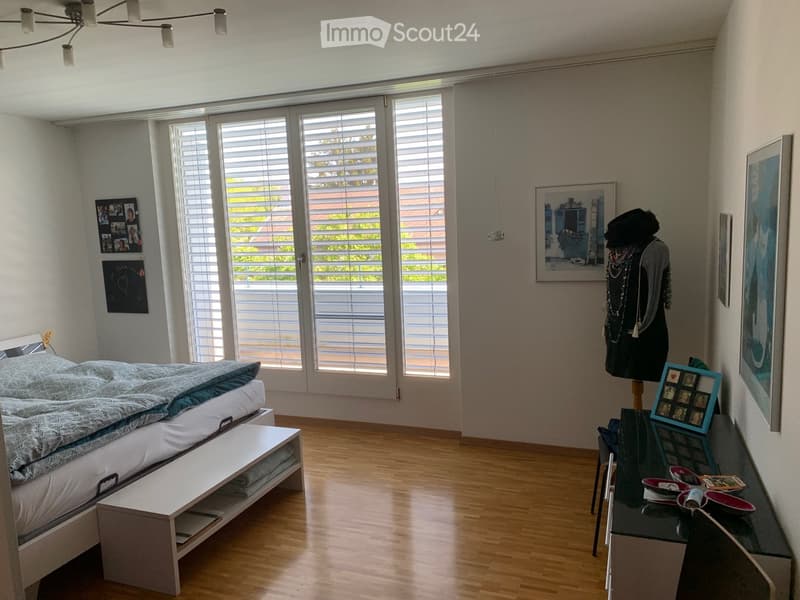 Bright bedroom with wooden flooring, large windows with blinds, a neatly made bed, a bench at the foot of the bed, a mirror, a chair, a small table with decorative items, framed pictures on the walls, and a mannequin with accessories.
