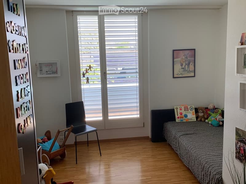 A cozy children's bedroom with a single bed, a chair, and various toys including a toy car and a toy horse. The room features wooden flooring, large windows with blinds, and decorative elements like picture frames and a name sign on the wall.