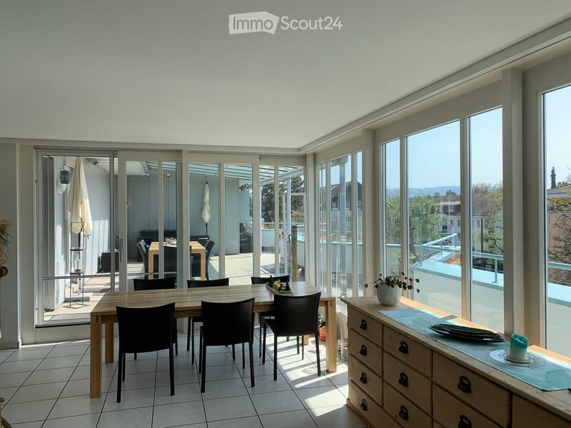 Bright, modern dining area with large windows and sliding glass doors leading to a patio. Features a wooden dining table with black chairs, a sideboard with a candle and vase, and an umbrella stand near the entrance.