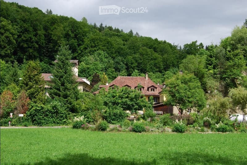 A two-story house with a red-tiled roof and yellow walls, surrounded by lush greenery and trees in a rural setting.