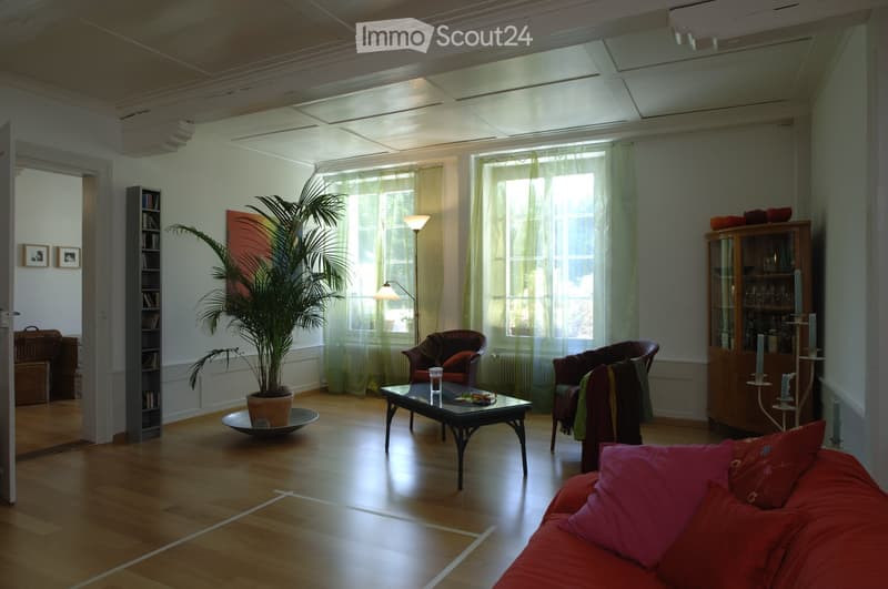 Bright living room with wooden flooring, large windows with sheer curtains, a red sofa, coffee table, armchairs, a potted plant, and a wooden cabinet with glass doors.