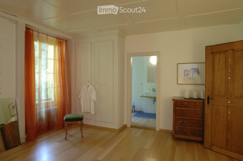 Spacious room with wooden flooring, large window with orange curtains, a green upholstered stool, a wooden wardrobe, a small dresser, and a partially visible bathroom with a sink and mirror.