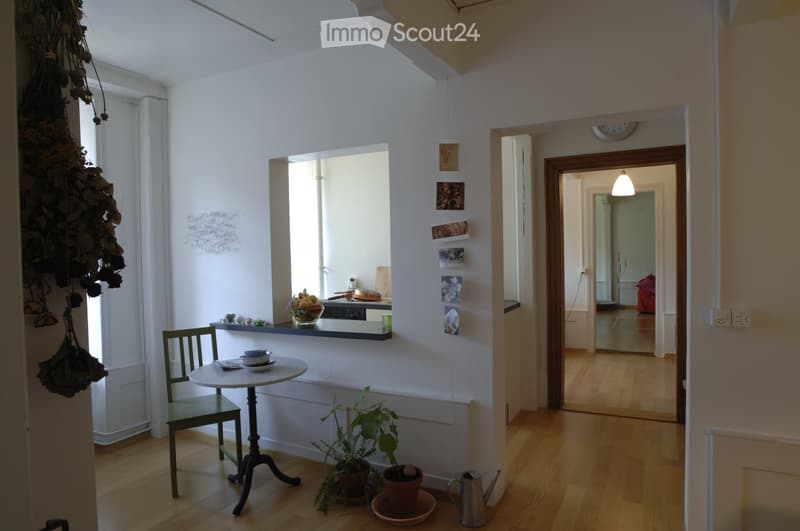 Bright hallway with wooden flooring, leading to a kitchen area and another room. Features include a small round table with a bowl and plates, a green chair, potted plants, and a hanging dried plant arrangement on the wall.