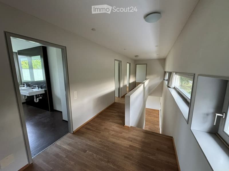 Empty hallway with wooden flooring, white walls, and a view into a bathroom area with a sink and mirror.