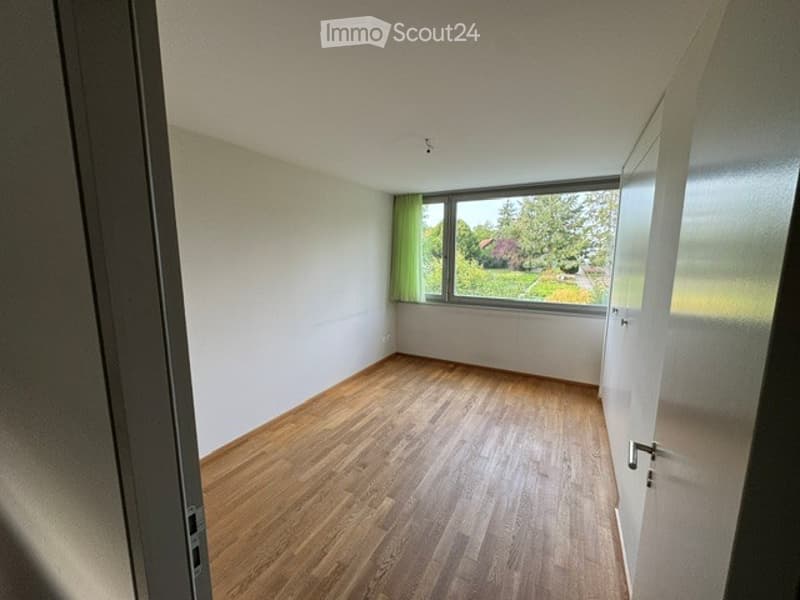 Empty room with wooden flooring, white walls, large window with green curtains, and a door visible on the right side.