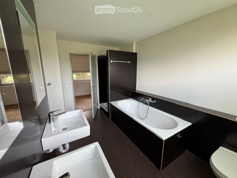 Modern bathroom with two sinks, a bathtub, and a toilet. The space features dark tiles and sleek fixtures.