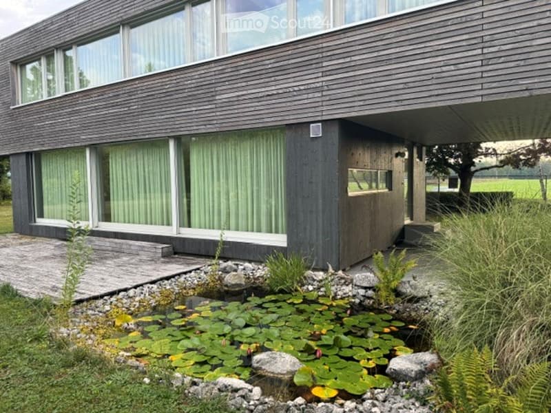 Modern two-story building with dark wooden facade and large windows, featuring a small pond with lily pads in the foreground.