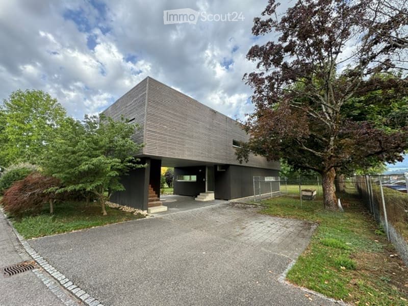 Modern two-story building with a raised ground floor, grey facade, and a small staircase leading to the entrance. The building is surrounded by trees and a paved driveway.