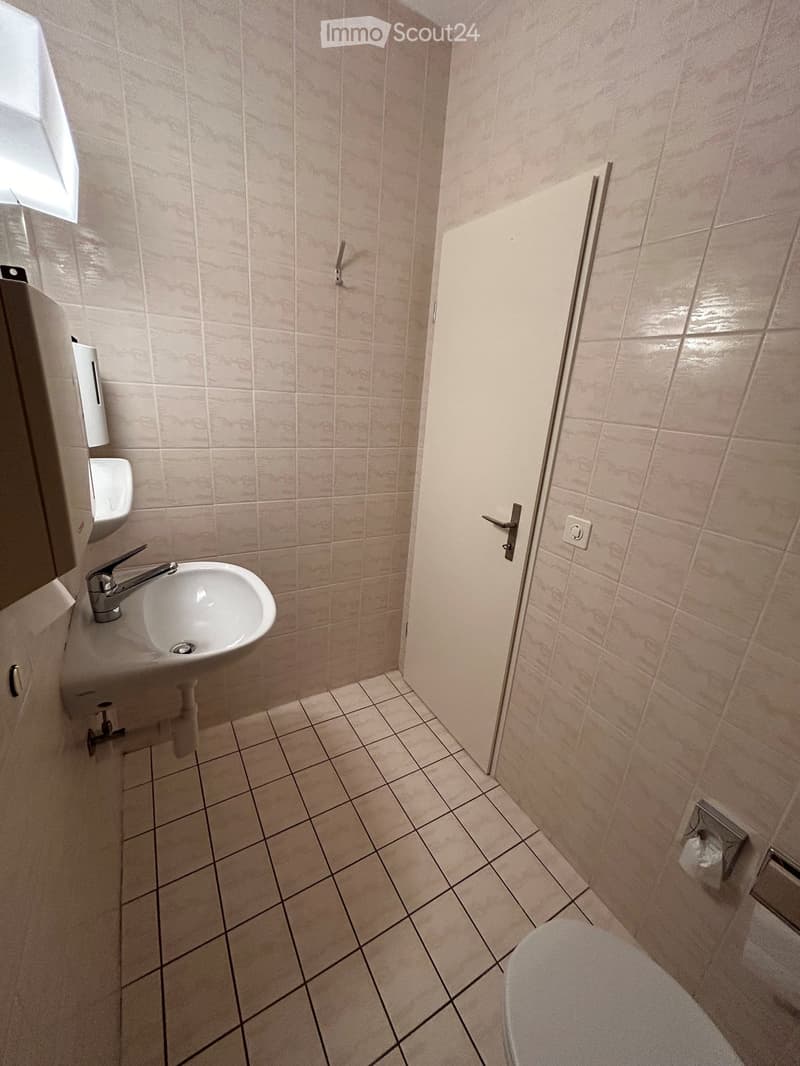 A small bathroom with tiled walls and floor, featuring a sink with a faucet, a toilet, and a door.