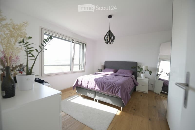 Modern bedroom with a grey bed, purple bedding, white nightstands, a large mirror, and potted plants. The room features wooden flooring and large windows allowing natural light.