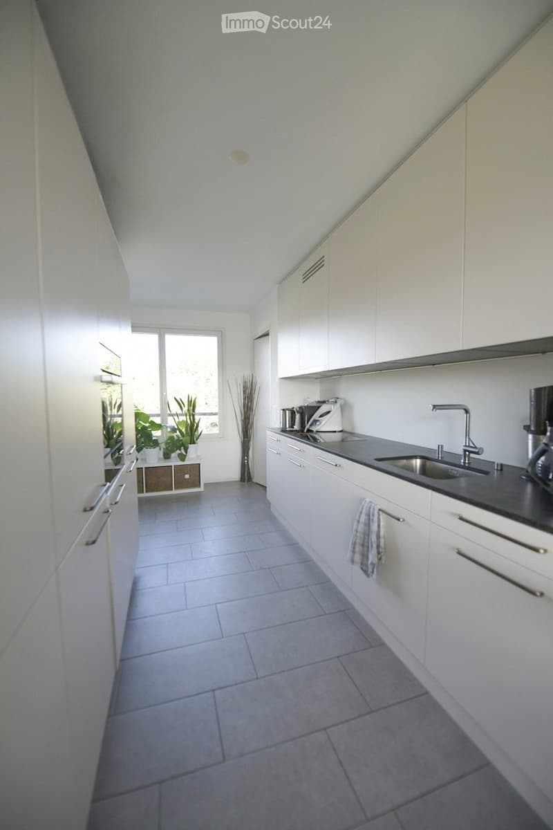 Modern kitchen with white cabinets, stainless steel sink, and tiled floor. Includes a coffee maker, oven, and a small decorative plant in the corner.