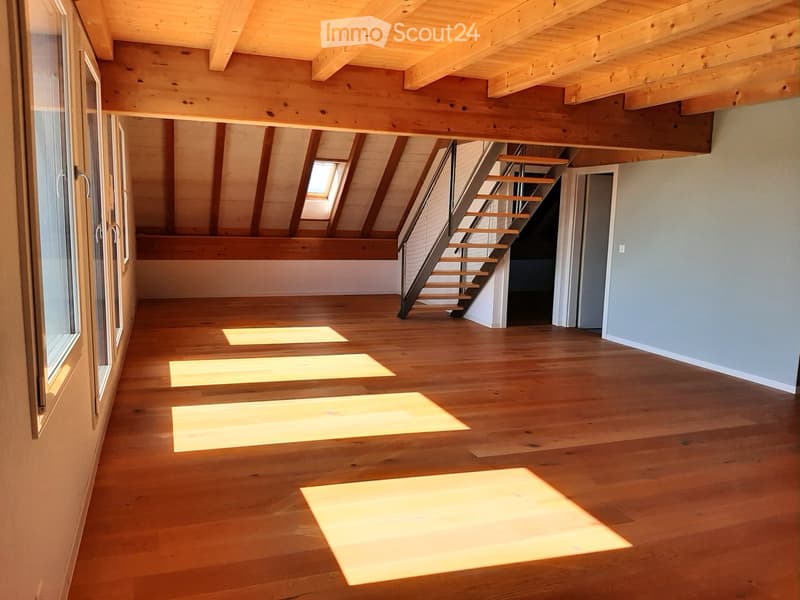 Bright, open attic space with wooden floors and exposed wooden beams. Features large windows and a staircase leading to another level.