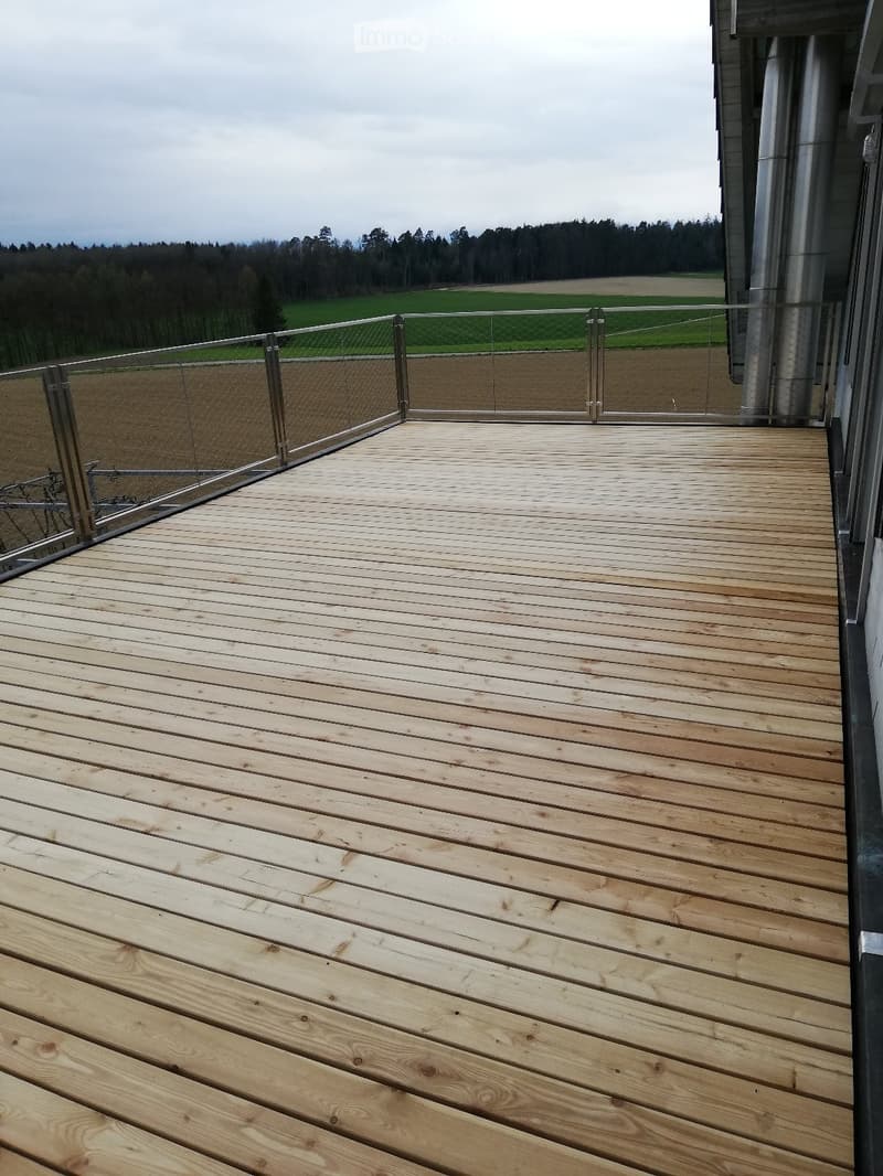 Wooden deck with glass railing, overlooking a green field and forest.