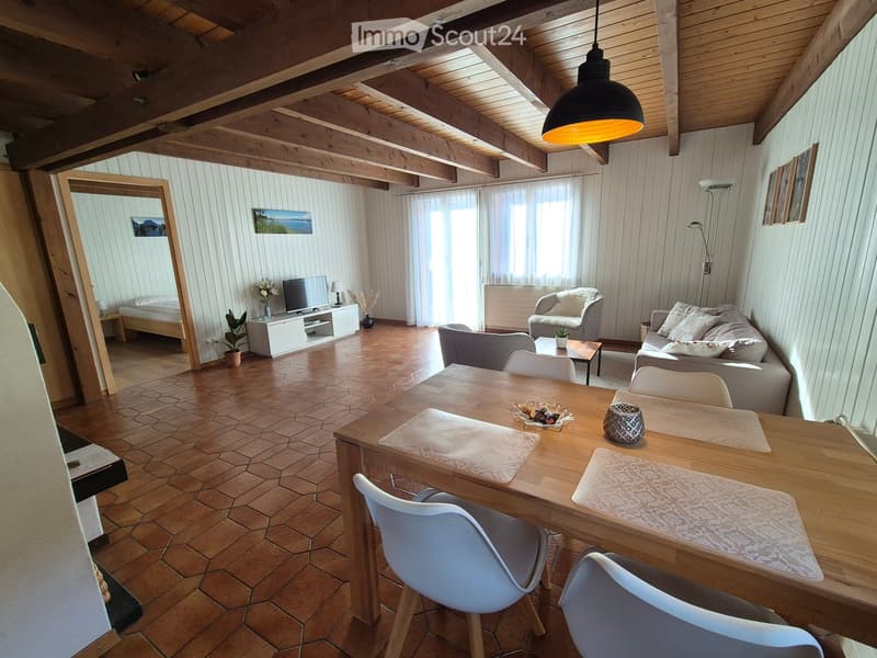 A cozy, well-lit living space with exposed wooden beams, a large wooden dining table with chairs, a sofa, a television, and a small bed visible in the background.
