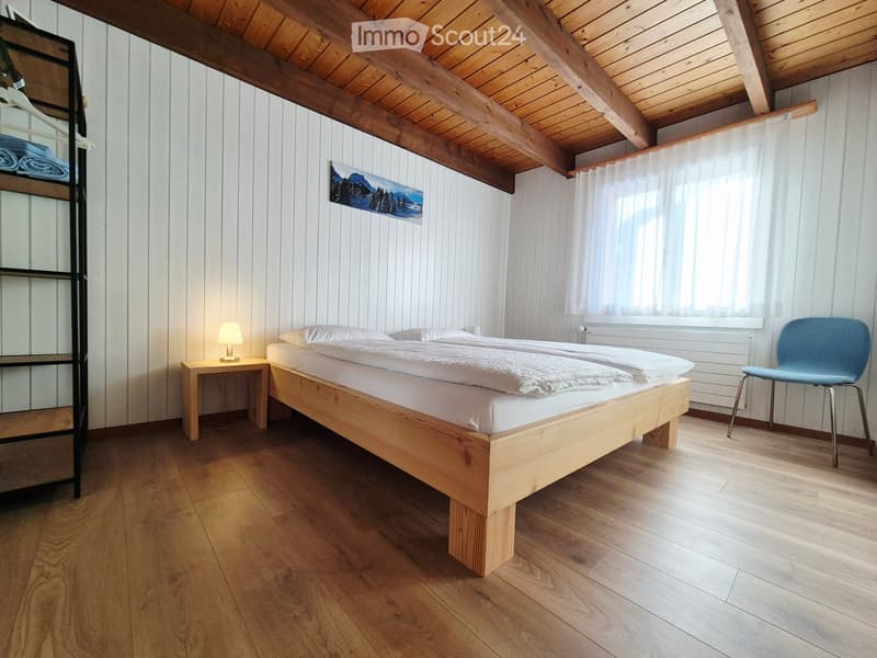 Cozy bedroom with wooden bed, nightstand, lamp, and a blue chair. Exposed wooden beams on the ceiling and a large window with white curtains.