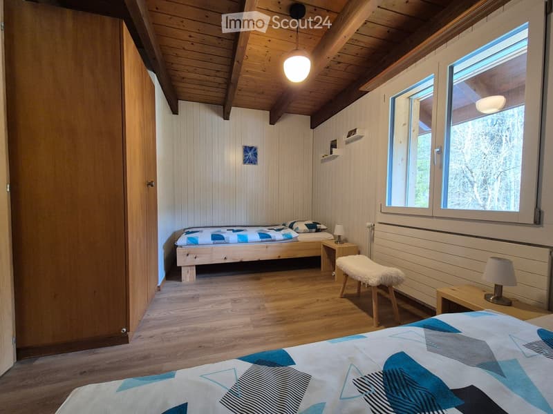 Cozy wooden bedroom with slanted ceiling, two beds with blue and white patterned bedding, small wooden table with lamp, and a fluffy white stool.