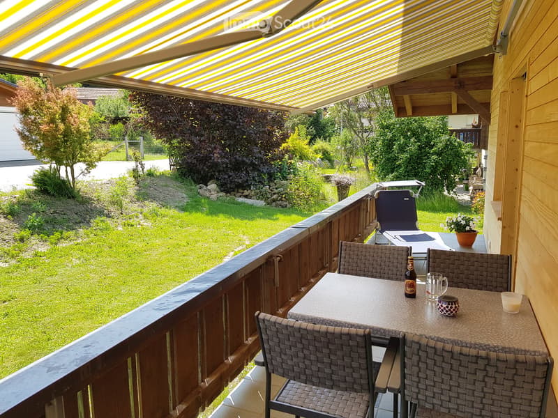 Balcony with a table, chairs, and a bottle on the table, overlooking a green garden with trees and a wooden fence.
