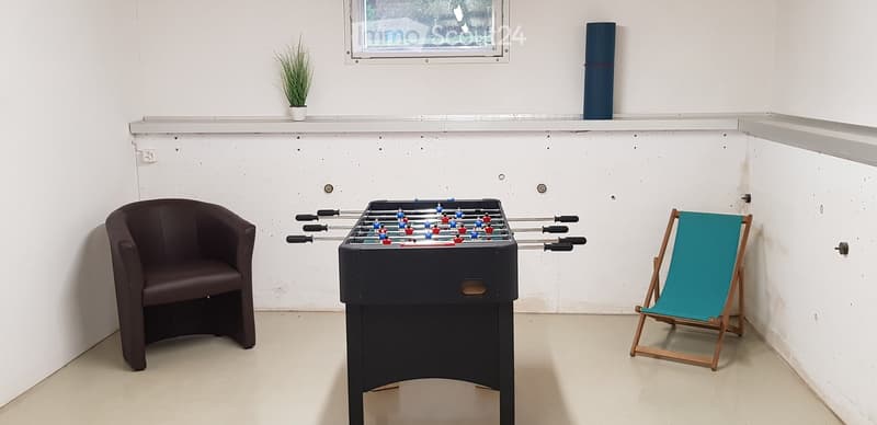 A small room with a foosball table, a dark brown leather chair, and a turquoise folding chair. There is a small potted plant on the windowsill and a dark blue cylindrical object on the shelf above the window.