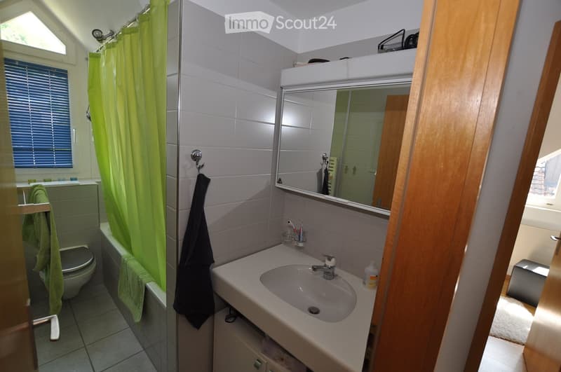 Modern bathroom with white tiles, a sink with a mirror above it, a toilet, and a shower area with a green curtain. A towel hangs on a rack, and a toothbrush holder is visible on the sink counter.