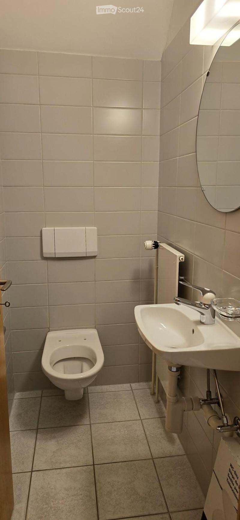 Small bathroom with beige tiled walls and floor, featuring a toilet, a sink with a modern faucet, and a round mirror above the sink.