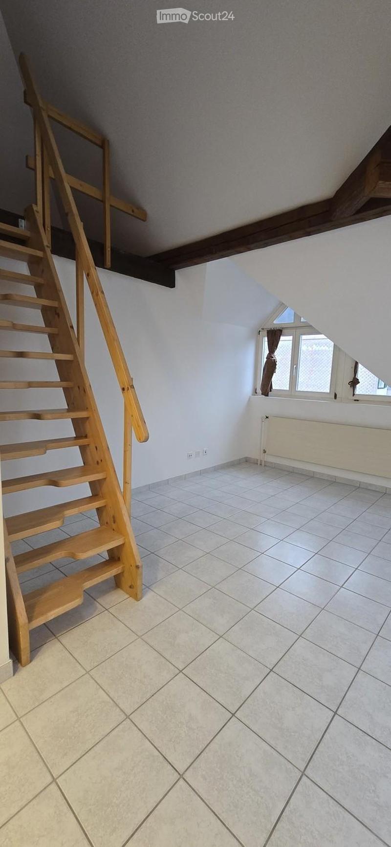 Empty attic room with wooden staircase, tiled floor, white walls, and a window with curtains.