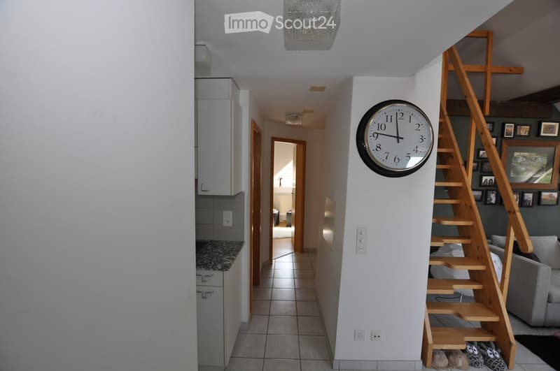 Modern hallway with a wooden staircase, wall clock, and visible kitchen and living room areas. The space features tiled flooring, white walls, and a chandelier hanging from the ceiling.