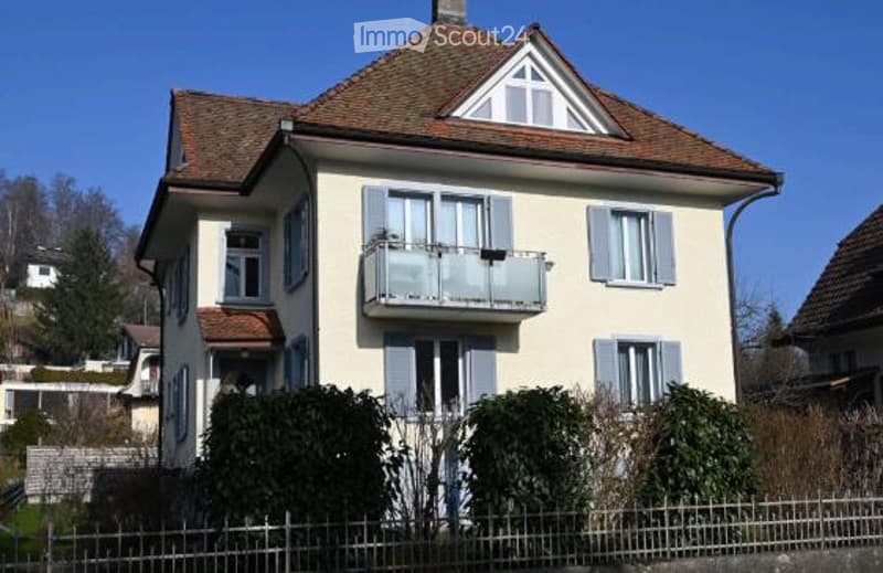 Two-story house with a cream-colored facade, gray shutters, and a balcony on the second floor. The roof is tiled with brown shingles, and the house is surrounded by greenery and a low fence.