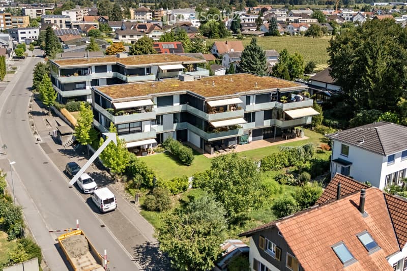 Aerial view of a modern multi-story residential building with a green roof, surrounded by trees and neighboring houses in a suburban neighborhood.