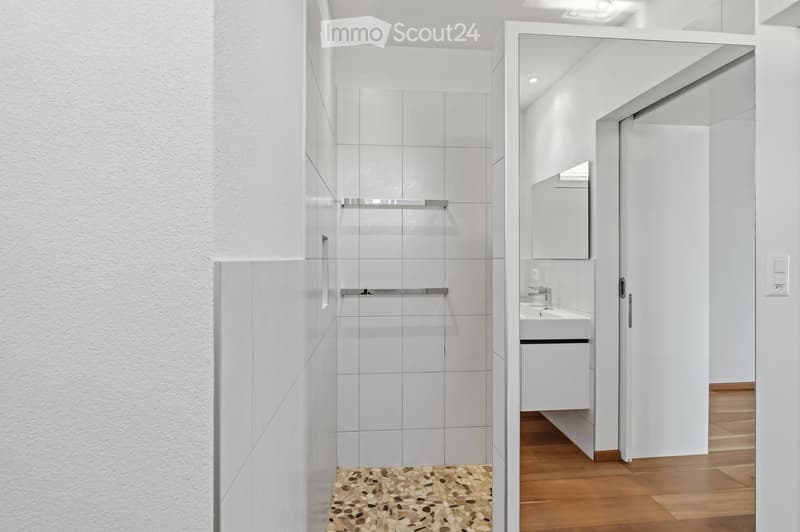 Modern bathroom with white tiles, a glass-enclosed shower, a sink with a mirror, and a wooden floor.