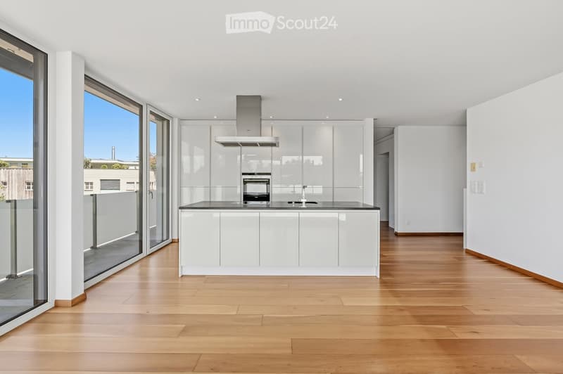 Modern kitchen with white cabinets, a central island, and a large window offering a view of the outside. The space features a sleek, minimalist design with a wooden floor and built-in appliances.